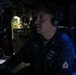 A U.S. Navy sailor is seated at a control station in a dimly lit operations room, wearing Sennheiser headphones and a navy blue uniform with 'U.S. NAVY' and a name tag reading 'BWANZ' visible. The environment appears to be a combat information center or bridge aboard a naval vessel, with multiple electronic displays and control panels visible. The sailor appears to be actively communicating or monitoring systems.