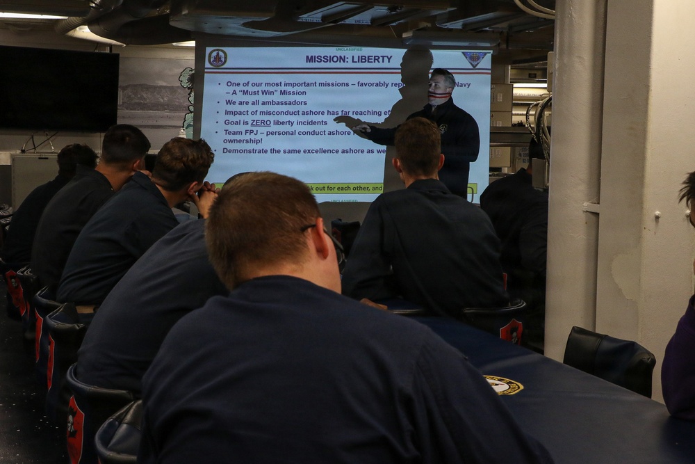 A military briefing is taking place in what appears to be a shipboard compartment, with a presenter standing in front of a projected slide titled 'MISSION: LIBERTY'. Several personnel in dark navy uniforms are seated facing the screen, attending the presentation. The slide contains bullet points about conduct ashore, liberty policies, and ambassador responsibilities.
