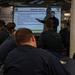 A military briefing is taking place in what appears to be a shipboard compartment, with a presenter standing in front of a projected slide titled 'MISSION: LIBERTY'. Several personnel in dark navy uniforms are seated facing the screen, attending the presentation. The slide contains bullet points about conduct ashore, liberty policies, and ambassador responsibilities.