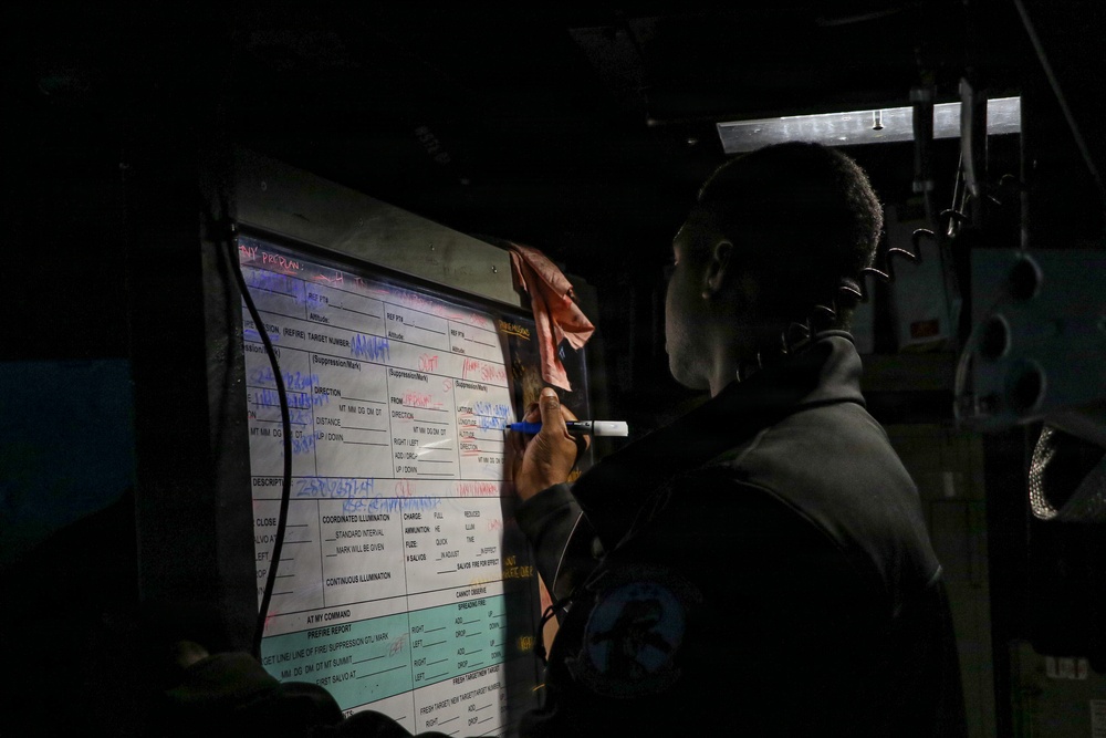 A person is writing on a military fire mission planning board in a low-light environment, likely inside a military vehicle or command post. The board contains pre-printed fire mission data forms with handwritten entries in multiple colors including red and blue marker. The scene appears to be a tactical military training or operational setting.