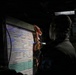 A person is writing on a military fire mission planning board in a low-light environment, likely inside a military vehicle or command post. The board contains pre-printed fire mission data forms with handwritten entries in multiple colors including red and blue marker. The scene appears to be a tactical military training or operational setting.