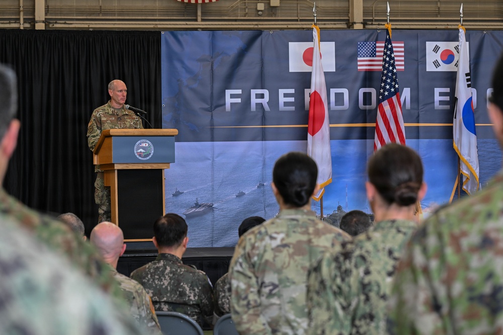A military officer in U.S. Army camouflage uniform speaks at a podium during a trilateral military ceremony or briefing. The backdrop displays the partial text 'FREEDOM E...' alongside the flags of Japan, the United States, and South Korea, suggesting a joint military exercise or event. An audience of mixed military personnel in various uniforms is seated and standing in attendance.