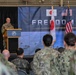 A military officer in U.S. Army camouflage uniform speaks at a podium during a trilateral military ceremony or briefing. The backdrop displays the partial text 'FREEDOM E...' alongside the flags of Japan, the United States, and South Korea, suggesting a joint military exercise or event. An audience of mixed military personnel in various uniforms is seated and standing in attendance.