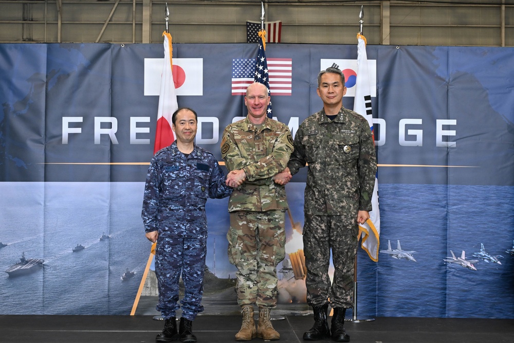 Three military officers representing Japan, the United States, and South Korea pose together at what appears to be the 'Freedom Edge' trilateral military exercise or event. The officers are shaking hands in front of a backdrop displaying the flags of all three nations along with military imagery. The setting appears to be inside a hangar or large military facility.