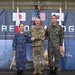 Three military officers representing Japan, the United States, and South Korea pose together at what appears to be the 'Freedom Edge' trilateral military exercise or event. The officers are shaking hands in front of a backdrop displaying the flags of all three nations along with military imagery. The setting appears to be inside a hangar or large military facility.