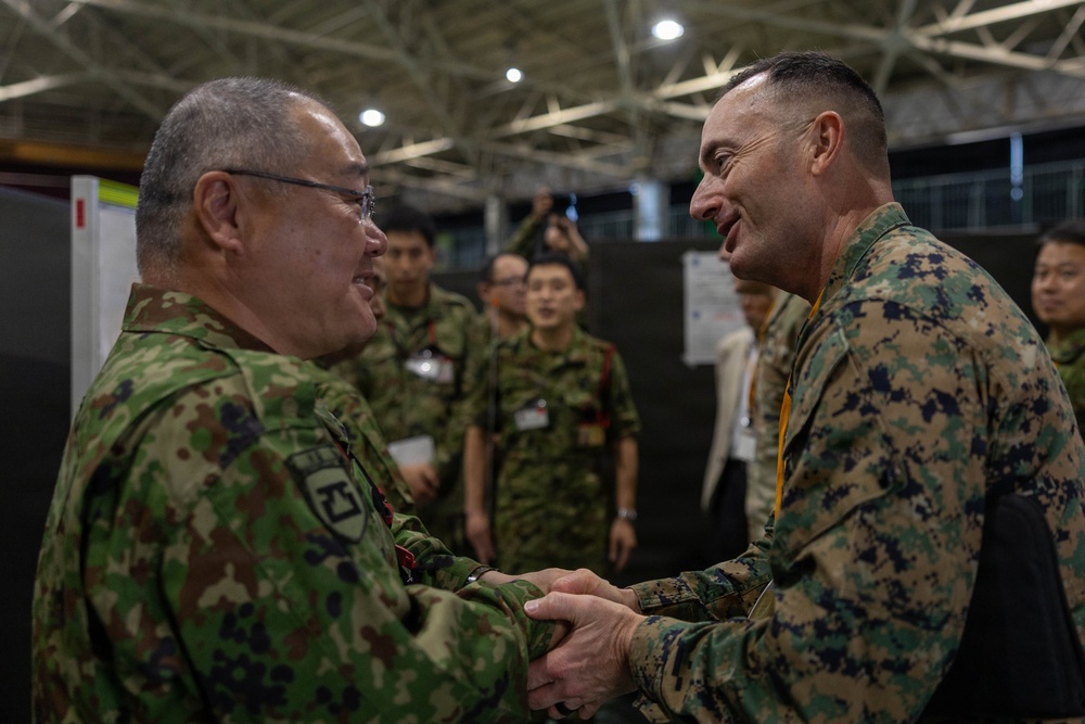Two military officers from different nations are shaking hands in what appears to be an indoor military exercise or conference facility. The officer on the left wears Japanese Ground Self-Defense Force (JGSDF) camouflage uniform, while the officer on the right wears United States Marine Corps (USMC) MARPAT digital camouflage. Both are smiling, suggesting a cordial bilateral military engagement or joint exercise event.