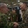 Two military officers from different nations are shaking hands in what appears to be an indoor military exercise or conference facility. The officer on the left wears Japanese Ground Self-Defense Force (JGSDF) camouflage uniform, while the officer on the right wears United States Marine Corps (USMC) MARPAT digital camouflage. Both are smiling, suggesting a cordial bilateral military engagement or joint exercise event.
