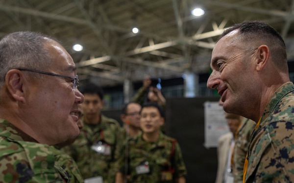 Two military officers from different nations are shaking hands in what appears to be an indoor military exercise or conference facility. The officer on the left wears Japanese Ground Self-Defense Force (JGSDF) camouflage uniform, while the officer on the right wears United States Marine Corps (USMC) MARPAT digital camouflage. Both are smiling, suggesting a cordial bilateral military engagement or joint exercise event.