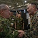 Two military officers from different nations are shaking hands in what appears to be an indoor military exercise or conference facility. The officer on the left wears Japanese Ground Self-Defense Force (JGSDF) camouflage uniform, while the officer on the right wears United States Marine Corps (USMC) MARPAT digital camouflage. Both are smiling, suggesting a cordial bilateral military engagement or joint exercise event.