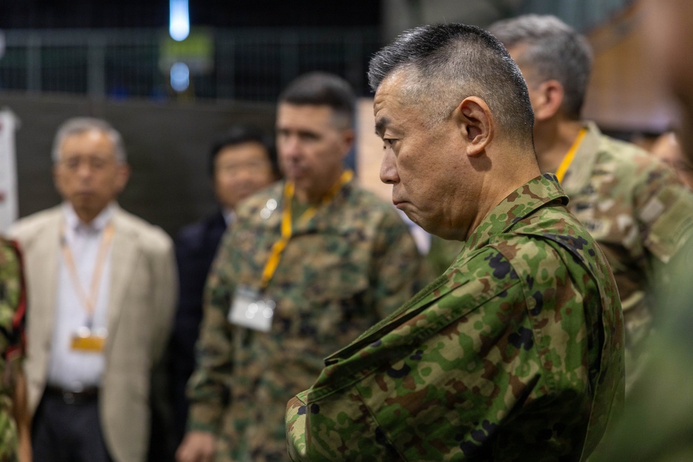 The image shows a group of military personnel and civilians gathered in what appears to be an indoor briefing or command center environment. The foreground features a senior officer in Japanese Ground Self-Defense Force (JGSDF) camouflage uniform with arms crossed, appearing attentive. Behind him are additional military personnel in mixed uniforms and civilians wearing lanyards with badges.