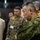 The image shows a group of military personnel and civilians gathered in what appears to be an indoor briefing or command center environment. The foreground features a senior officer in Japanese Ground Self-Defense Force (JGSDF) camouflage uniform with arms crossed, appearing attentive. Behind him are additional military personnel in mixed uniforms and civilians wearing lanyards with badges.