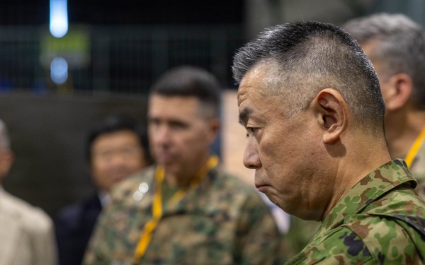 The image shows a group of military personnel and civilians gathered in what appears to be an indoor briefing or command center environment. The foreground features a senior officer in Japanese Ground Self-Defense Force (JGSDF) camouflage uniform with arms crossed, appearing attentive. Behind him are additional military personnel in mixed uniforms and civilians wearing lanyards with badges.