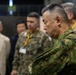 The image shows a group of military personnel and civilians gathered in what appears to be an indoor briefing or command center environment. The foreground features a senior officer in Japanese Ground Self-Defense Force (JGSDF) camouflage uniform with arms crossed, appearing attentive. Behind him are additional military personnel in mixed uniforms and civilians wearing lanyards with badges.