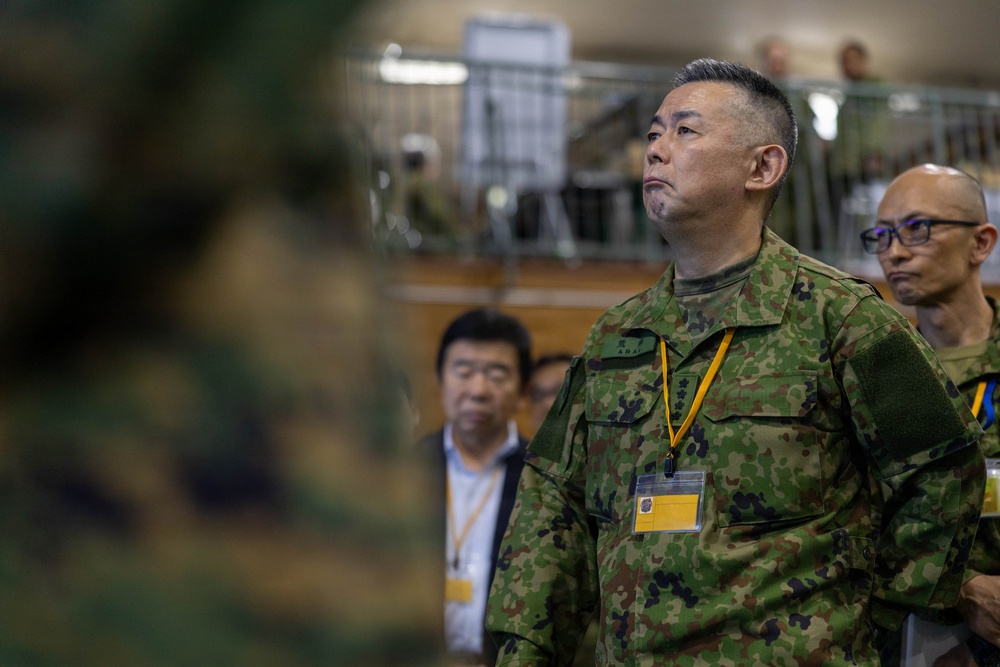 The image shows military personnel and civilians in what appears to be an indoor facility, likely during a briefing or inspection event. The central figure is wearing Japanese Ground Self-Defense Force (JGSDF) camouflage uniform with a yellow lanyard and identification badge. Several other individuals in both military and civilian attire are visible in the background.