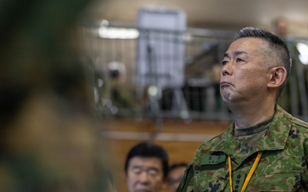 The image shows military personnel and civilians in what appears to be an indoor facility, likely during a briefing or inspection event. The central figure is wearing Japanese Ground Self-Defense Force (JGSDF) camouflage uniform with a yellow lanyard and identification badge. Several other individuals in both military and civilian attire are visible in the background.