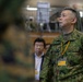 The image shows military personnel and civilians in what appears to be an indoor facility, likely during a briefing or inspection event. The central figure is wearing Japanese Ground Self-Defense Force (JGSDF) camouflage uniform with a yellow lanyard and identification badge. Several other individuals in both military and civilian attire are visible in the background.