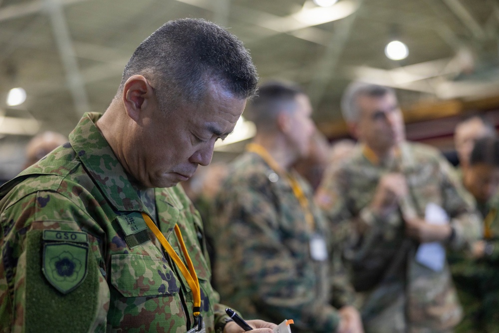A military officer in Japanese Ground Self-Defense Force (JGSDF) camouflage uniform is focused on writing notes, wearing a yellow lanyard and a patch reading 'GSO' on his left shoulder. He is surrounded by other military personnel in various camouflage uniforms in what appears to be an indoor military facility or hangar. The setting suggests a multinational military exercise, briefing, or coordination event.