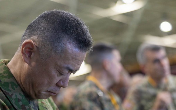 A military officer in Japanese Ground Self-Defense Force (JGSDF) camouflage uniform is focused on writing notes, wearing a yellow lanyard and a patch reading 'GSO' on his left shoulder. He is surrounded by other military personnel in various camouflage uniforms in what appears to be an indoor military facility or hangar. The setting suggests a multinational military exercise, briefing, or coordination event.
