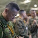 A military officer in Japanese Ground Self-Defense Force (JGSDF) camouflage uniform is focused on writing notes, wearing a yellow lanyard and a patch reading 'GSO' on his left shoulder. He is surrounded by other military personnel in various camouflage uniforms in what appears to be an indoor military facility or hangar. The setting suggests a multinational military exercise, briefing, or coordination event.