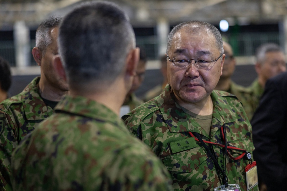 The image shows military personnel in Japanese Ground Self-Defense Force (JGSDF) camouflage uniforms engaged in conversation. The central figure, identified by a name tape reading 'TORIUMI' in both Japanese characters (鳥海) and Roman letters, is wearing glasses and a lanyard with an ID badge. The setting appears to be an indoor military facility or event with multiple personnel present.