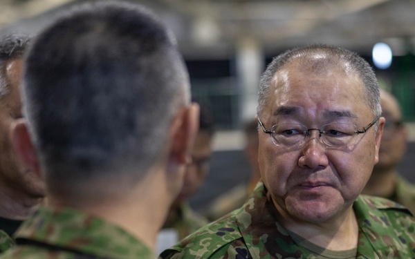 The image shows military personnel in Japanese Ground Self-Defense Force (JGSDF) camouflage uniforms engaged in conversation. The central figure, identified by a name tape reading 'TORIUMI' in both Japanese characters (鳥海) and Roman letters, is wearing glasses and a lanyard with an ID badge. The setting appears to be an indoor military facility or event with multiple personnel present.