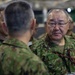 The image shows military personnel in Japanese Ground Self-Defense Force (JGSDF) camouflage uniforms engaged in conversation. The central figure, identified by a name tape reading 'TORIUMI' in both Japanese characters (鳥海) and Roman letters, is wearing glasses and a lanyard with an ID badge. The setting appears to be an indoor military facility or event with multiple personnel present.