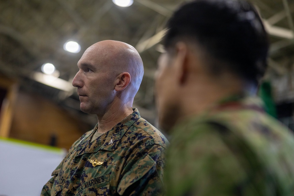 A U.S. Marine in MARPAT camouflage uniform is shown in profile view inside what appears to be a large indoor facility, likely a hangar or gymnasium. He is facing a second individual in the foreground who is out of focus and appears to be wearing a different military uniform. The setting suggests a formal or professional military interaction or briefing.