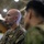 A U.S. Marine in MARPAT camouflage uniform is shown in profile view inside what appears to be a large indoor facility, likely a hangar or gymnasium. He is facing a second individual in the foreground who is out of focus and appears to be wearing a different military uniform. The setting suggests a formal or professional military interaction or briefing.