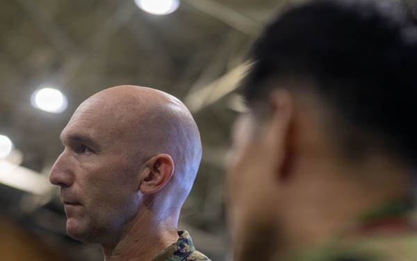 A U.S. Marine in MARPAT camouflage uniform is shown in profile view inside what appears to be a large indoor facility, likely a hangar or gymnasium. He is facing a second individual in the foreground who is out of focus and appears to be wearing a different military uniform. The setting suggests a formal or professional military interaction or briefing.