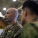 A U.S. Marine in MARPAT camouflage uniform is shown in profile view inside what appears to be a large indoor facility, likely a hangar or gymnasium. He is facing a second individual in the foreground who is out of focus and appears to be wearing a different military uniform. The setting suggests a formal or professional military interaction or briefing.