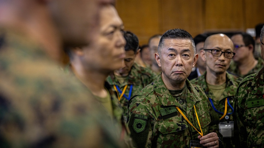 A group of military personnel in camouflage uniforms are gathered in what appears to be an indoor formal or briefing setting. The uniforms and insignia are consistent with the Japan Ground Self-Defense Force (JGSDF), with visible name tags and unit patches. Personnel are wearing lanyards with identification badges in blue and yellow colors.