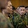 A group of military personnel in camouflage uniforms are gathered in what appears to be an indoor formal or briefing setting. The uniforms and insignia are consistent with the Japan Ground Self-Defense Force (JGSDF), with visible name tags and unit patches. Personnel are wearing lanyards with identification badges in blue and yellow colors.