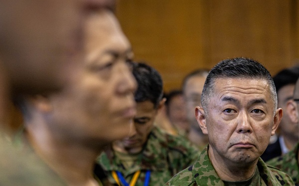 A group of military personnel in camouflage uniforms are gathered in what appears to be an indoor formal or briefing setting. The uniforms and insignia are consistent with the Japan Ground Self-Defense Force (JGSDF), with visible name tags and unit patches. Personnel are wearing lanyards with identification badges in blue and yellow colors.
