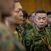 A group of military personnel in camouflage uniforms are gathered in what appears to be an indoor formal or briefing setting. The uniforms and insignia are consistent with the Japan Ground Self-Defense Force (JGSDF), with visible name tags and unit patches. Personnel are wearing lanyards with identification badges in blue and yellow colors.