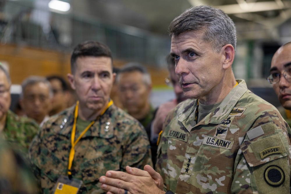 A U.S. Army officer with the name tape 'McFARLANE' and two-star general rank insignia is speaking to a group in what appears to be a military hangar or facility. He is wearing OCP (Operational Camouflage Pattern) uniform with a Ranger tab and U.S. Army identification. A U.S. Marine Corps officer in MARPAT uniform is visible in the background alongside other military personnel.