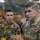 A U.S. Army officer with the name tape 'McFARLANE' and two-star general rank insignia is speaking to a group in what appears to be a military hangar or facility. He is wearing OCP (Operational Camouflage Pattern) uniform with a Ranger tab and U.S. Army identification. A U.S. Marine Corps officer in MARPAT uniform is visible in the background alongside other military personnel.