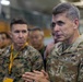 A U.S. Army officer with the name tape 'McFARLANE' and two-star general rank insignia is speaking to a group in what appears to be a military hangar or facility. He is wearing OCP (Operational Camouflage Pattern) uniform with a Ranger tab and U.S. Army identification. A U.S. Marine Corps officer in MARPAT uniform is visible in the background alongside other military personnel.