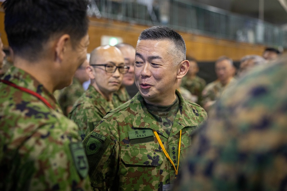The image shows a group of military personnel in what appears to be an indoor briefing or meeting environment. The central figure is wearing Japanese Ground Self-Defense Force (JGSDF) camouflage uniform with a name tape reading 'ARAI' and rank insignia visible. Personnel from what appears to be both JGSDF and U.S. Marine Corps are present, suggesting a joint military exercise or coordination event.