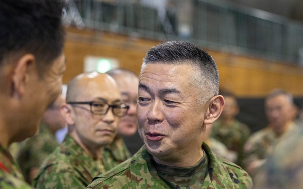 The image shows a group of military personnel in what appears to be an indoor briefing or meeting environment. The central figure is wearing Japanese Ground Self-Defense Force (JGSDF) camouflage uniform with a name tape reading 'ARAI' and rank insignia visible. Personnel from what appears to be both JGSDF and U.S. Marine Corps are present, suggesting a joint military exercise or coordination event.