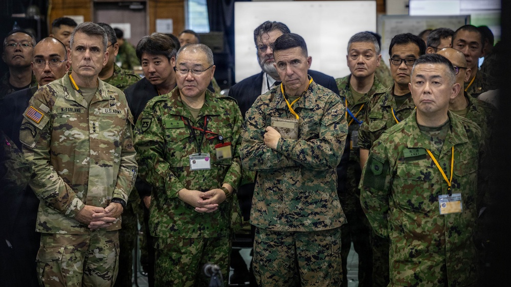 A group of military personnel from what appears to be the United States Army, U.S. Marines, and Japanese Self-Defense Forces are gathered together in an indoor command or briefing facility. The individuals are standing attentively, likely observing a briefing or presentation. The mix of U.S. and Japanese uniforms suggests a joint military exercise or coordination meeting.