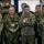A group of military personnel from what appears to be the United States Army, U.S. Marines, and Japanese Self-Defense Forces are gathered together in an indoor command or briefing facility. The individuals are standing attentively, likely observing a briefing or presentation. The mix of U.S. and Japanese uniforms suggests a joint military exercise or coordination meeting.
