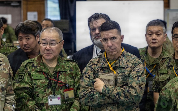 A group of military personnel from what appears to be the United States Army, U.S. Marines, and Japanese Self-Defense Forces are gathered together in an indoor command or briefing facility. The individuals are standing attentively, likely observing a briefing or presentation. The mix of U.S. and Japanese uniforms suggests a joint military exercise or coordination meeting.