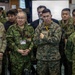 A group of military personnel from what appears to be the United States Army, U.S. Marines, and Japanese Self-Defense Forces are gathered together in an indoor command or briefing facility. The individuals are standing attentively, likely observing a briefing or presentation. The mix of U.S. and Japanese uniforms suggests a joint military exercise or coordination meeting.