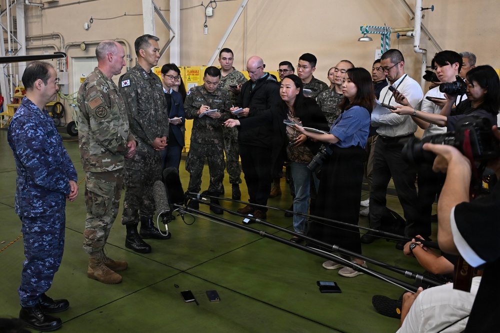 A military press briefing or media availability is taking place inside what appears to be a military hangar or facility. Three military personnel in different uniforms — including U.S. Army camouflage, a South Korean military uniform with a visible ROK flag patch, and a blue digital camouflage uniform — are facing a group of journalists and media personnel. Reporters are holding microphones, boom poles, cameras, and notepads, suggesting an official military press event.