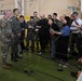 A military press briefing or media availability is taking place inside what appears to be a military hangar or facility. Three military personnel in different uniforms — including U.S. Army camouflage, a South Korean military uniform with a visible ROK flag patch, and a blue digital camouflage uniform — are facing a group of journalists and media personnel. Reporters are holding microphones, boom poles, cameras, and notepads, suggesting an official military press event.