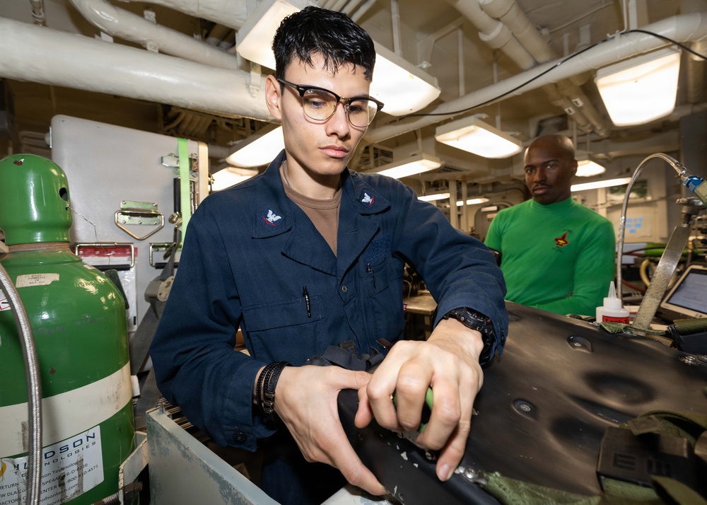 Two military personnel, likely U.S. Navy sailors, are working in what appears to be a shipboard maintenance or repair shop. The foreground sailor in navy blue coveralls is focused on working with a dark-colored component or equipment piece, while a second sailor in a green shirt observes or works in the background. The environment shows an industrial interior consistent with a naval vessel workspace.