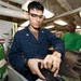 Two military personnel, likely U.S. Navy sailors, are working in what appears to be a shipboard maintenance or repair shop. The foreground sailor in navy blue coveralls is focused on working with a dark-colored component or equipment piece, while a second sailor in a green shirt observes or works in the background. The environment shows an industrial interior consistent with a naval vessel workspace.