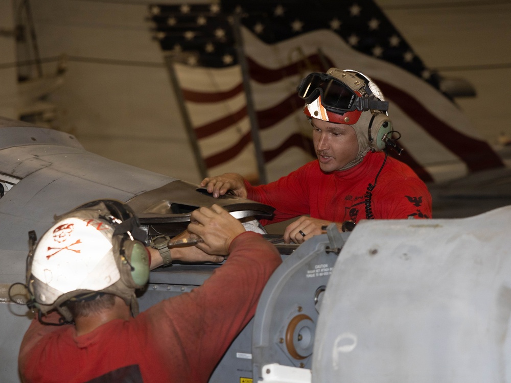 Two military personnel wearing red shirts and cranial helmets with hearing protection are performing maintenance work on an aircraft in what appears to be a hangar bay. A large American flag is prominently displayed in the background, suggesting this is likely aboard a U.S. Navy aircraft carrier. The workers are accessing an aircraft panel or compartment, consistent with aviation ordnance or maintenance crew duties.