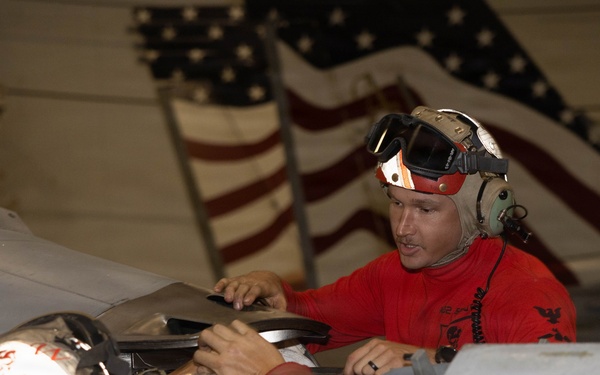 Two military personnel wearing red shirts and cranial helmets with hearing protection are performing maintenance work on an aircraft in what appears to be a hangar bay. A large American flag is prominently displayed in the background, suggesting this is likely aboard a U.S. Navy aircraft carrier. The workers are accessing an aircraft panel or compartment, consistent with aviation ordnance or maintenance crew duties.