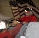 Two military personnel wearing red shirts and cranial helmets with hearing protection are performing maintenance work on an aircraft in what appears to be a hangar bay. A large American flag is prominently displayed in the background, suggesting this is likely aboard a U.S. Navy aircraft carrier. The workers are accessing an aircraft panel or compartment, consistent with aviation ordnance or maintenance crew duties.